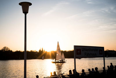 Silhouette people on street by lake against sky during sunset