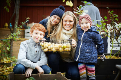 Portrait of smiling mother with potato basket crouching by children at yard