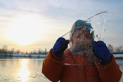 Woman holding ice over face while standing by lake against sky during winter
