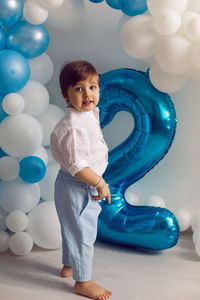 Baby boy in blue pants and shirt standing on the floor in blue and white balloons