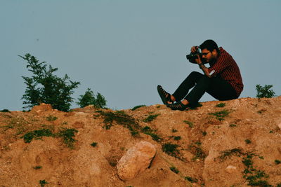 Full length of young man photographing through camera while sitting on mountain against sky