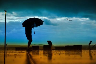 Silhouette man holding umbrella while walking at beach against cloudy sky during monsoon