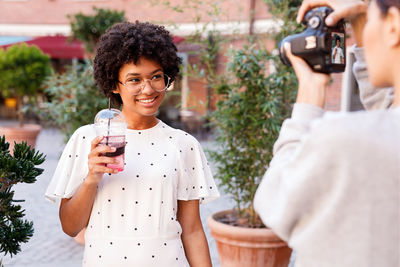 Girl photographing friend while holding drinking glass against plants