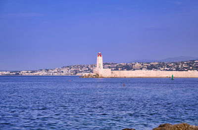 Lighthouse amidst sea and buildings against clear blue sky