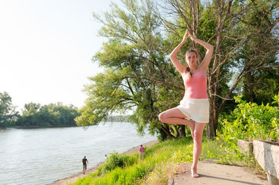 Full length of young woman doing yoga by river on sunny day