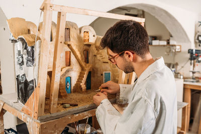 Side view of craftsman making model of facade of clay house and creating small wooden door while working in art workshop