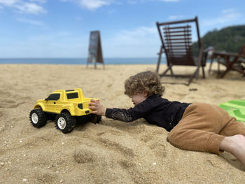 Blonde child lying on the fine sand playing with her toy yellow pickup truck on a sunny summer day