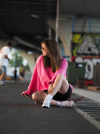 Young beautiful stylish girl skater sits on a skateboard in a modern skate park.