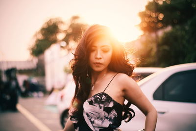 Beautiful young woman standing against cars during sunset