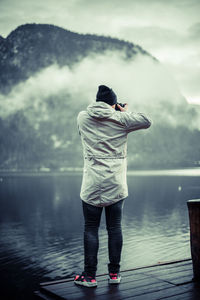 Full length rear view of man photographing by lake against mountain during foggy weather