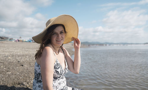 Woman sitting alone at the edge of the beach looking at the camera, holding her hat with her hand