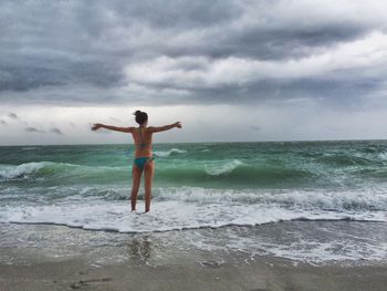 Rear view of sensuous young woman standing on sea shore against cloudy sky in stormy weather