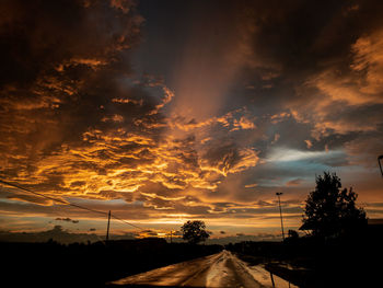 Road by silhouette trees against dramatic sky during sunset