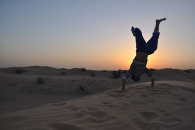 Man practicing handstand on desert against sky during sunset
