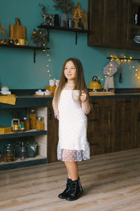 A little girl in a white dress holds a cup of tea in the kitchen, decorated for christmas 