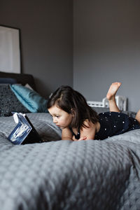 Full length of cute girl watching digital tablet while lying on bed at home