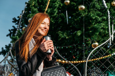 Smiling young woman looking away while holding coffee cup against trees