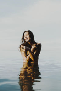 Cheerful young woman applying mud on hands while standing in lake against sky
