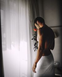 Shirtless young man holding rose while standing by curtain at home