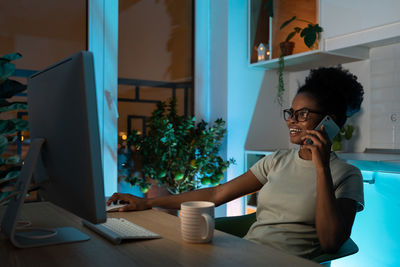Positive multiracial woman in glasses talking on smartphone, working on desktop pc late at night