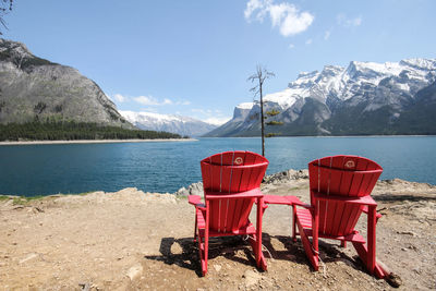 Empty red adirondack chairs at lakeshore against snowcapped mountains