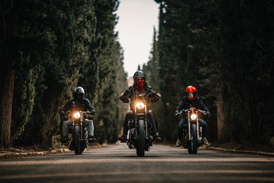 Group of bikers in black leather jackets and helmets riding powerful motorcycles on asphalt road leading between green forest in countryside