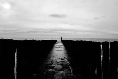 View of wooden walkway leading towards water against sky