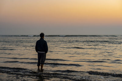 Rear view of man standing on beach against sky during sunset