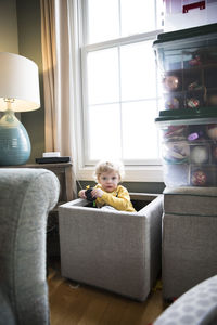 Camera aware toddler sits in toy box in living room in front of window