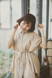 Smiling young woman listening music while standing by window at home