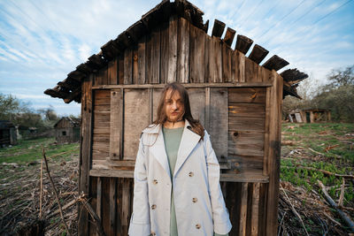 Beautiful girl with long hair in a grey trench coat next to an old wooden house outdoors in spring