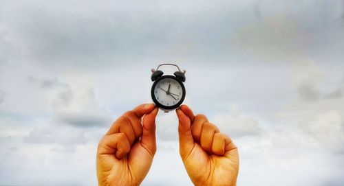 Close-up of cropped hands holding alarm clock against cloudy sky