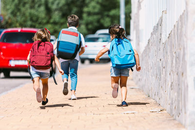 Back view of anonymous schoolkids with backpacks running on tiled walkway in sunny town on blurred background