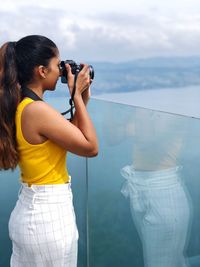 Young woman photographing while standing on sea against sky