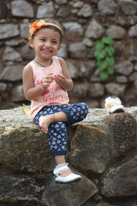 Full length of cute smiling girl looking away while sitting on stone wall
