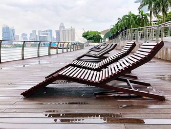 Wooden bench on pier by river against sky in city