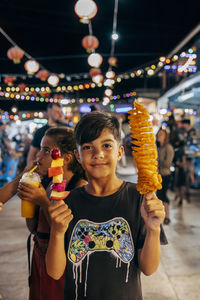 Portrait of smiling boy holding tornado potato fries and fruit kebab at food market