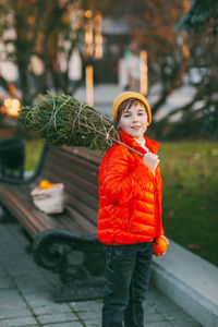 A cute boy in an orange jacket and hat walks through the park with a christmas tree on his shoulder. 