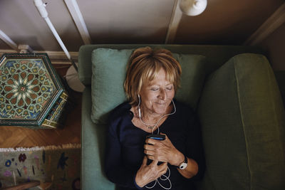 Smiling senior woman with smart phone listening music on sofa at home