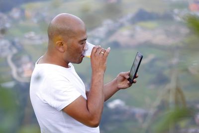 Side view of bald man drinking while using phone