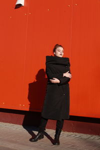 Fashionable teenage girl wearing black warm clothing standing against red wall during sunny day