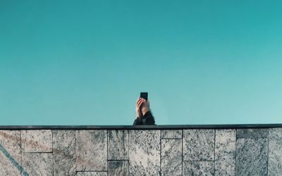 Man photographing woman standing on retaining wall against clear sky