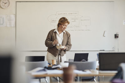 Female teacher holding lunch box while looking at laptop in classroom