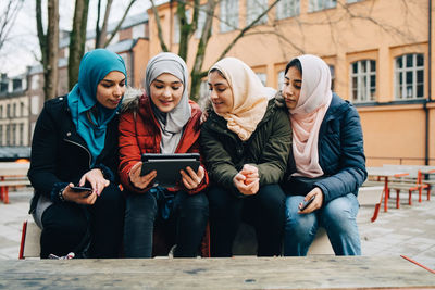 Female muslim friends sitting on bench sharing digital tablet in city