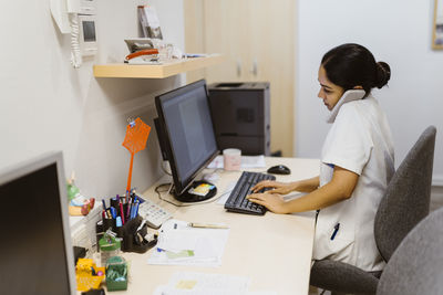 Side view of female doctor working on computer while talking through smart phone at clinic