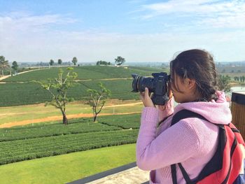 Side view of young woman photographing while standing against sky