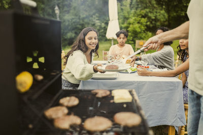 Midsection of father serving grilled patty to smiling daughter sitting on table in back yard