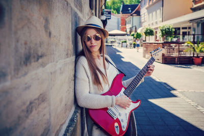 Portrait of young woman playing guitar while standing by wall