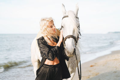 Young blonde woman in black clothes riding white horse on seascape background