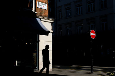 Silhouette man walking on street corner with sun against buildings 